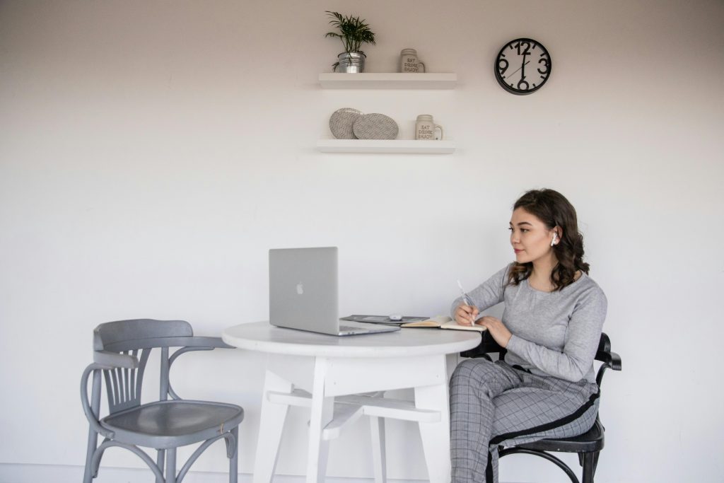 woman with earbuds looking at a laptop screen while taking notes in a notebook